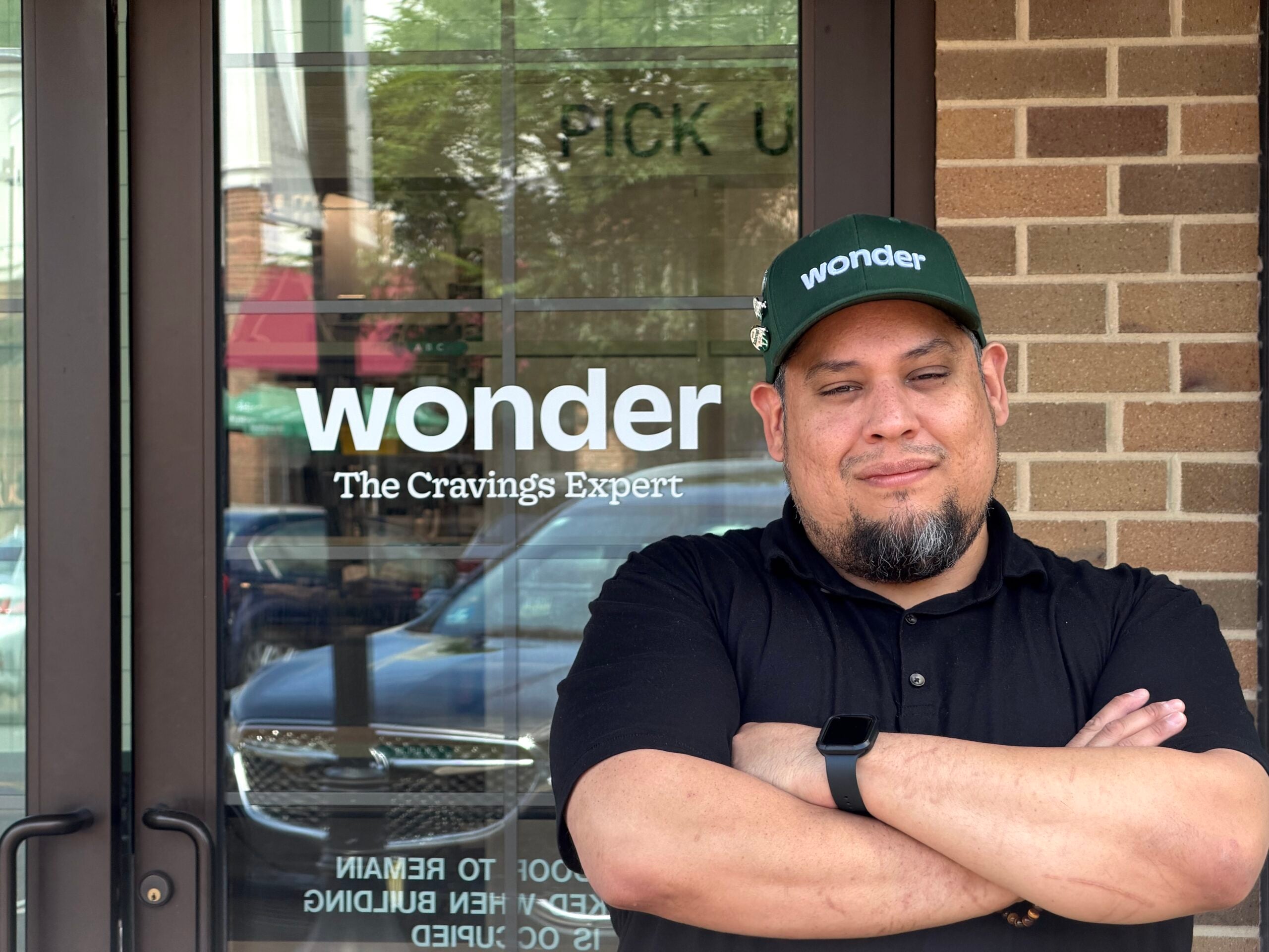 Man wearing a green Wonder hat and black shirt stands in front of a glass door.